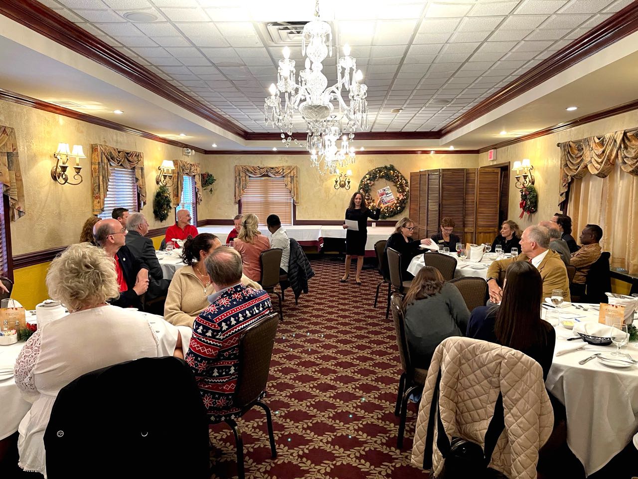 Group of people seated at tables listening to a woman speak at the front of the room.